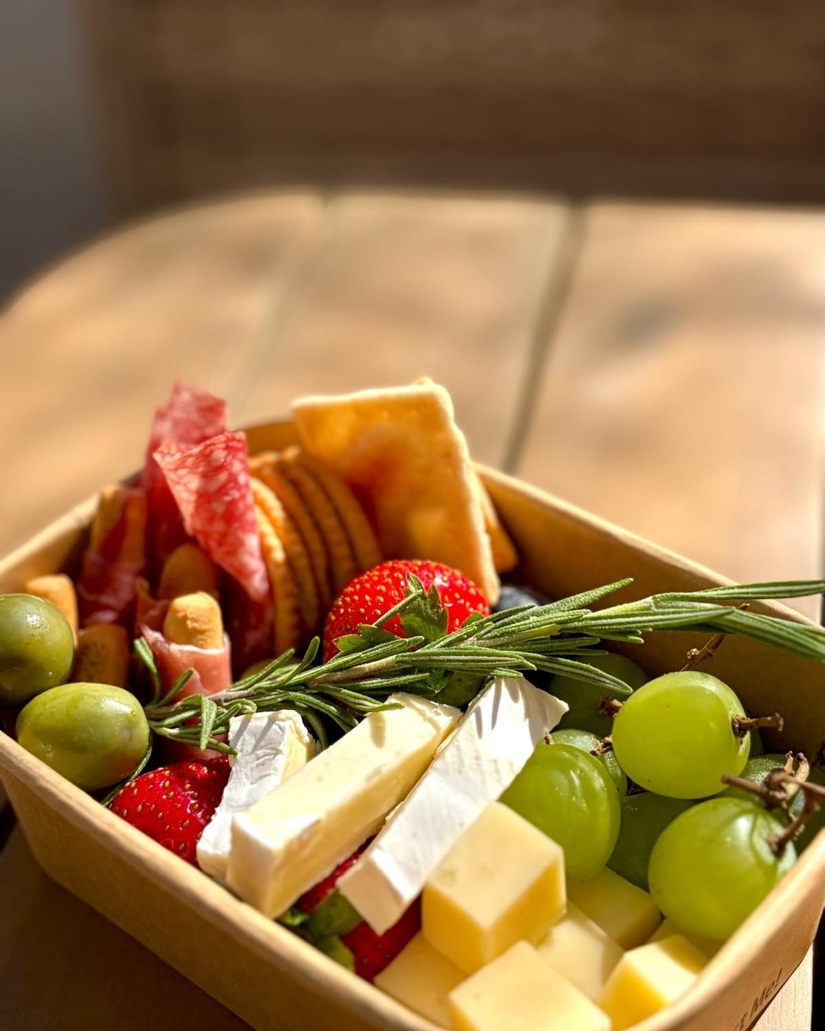 An open graze box of salami, brie, cheddar, grapes, strawberries and crackers with a sprig of rosemary