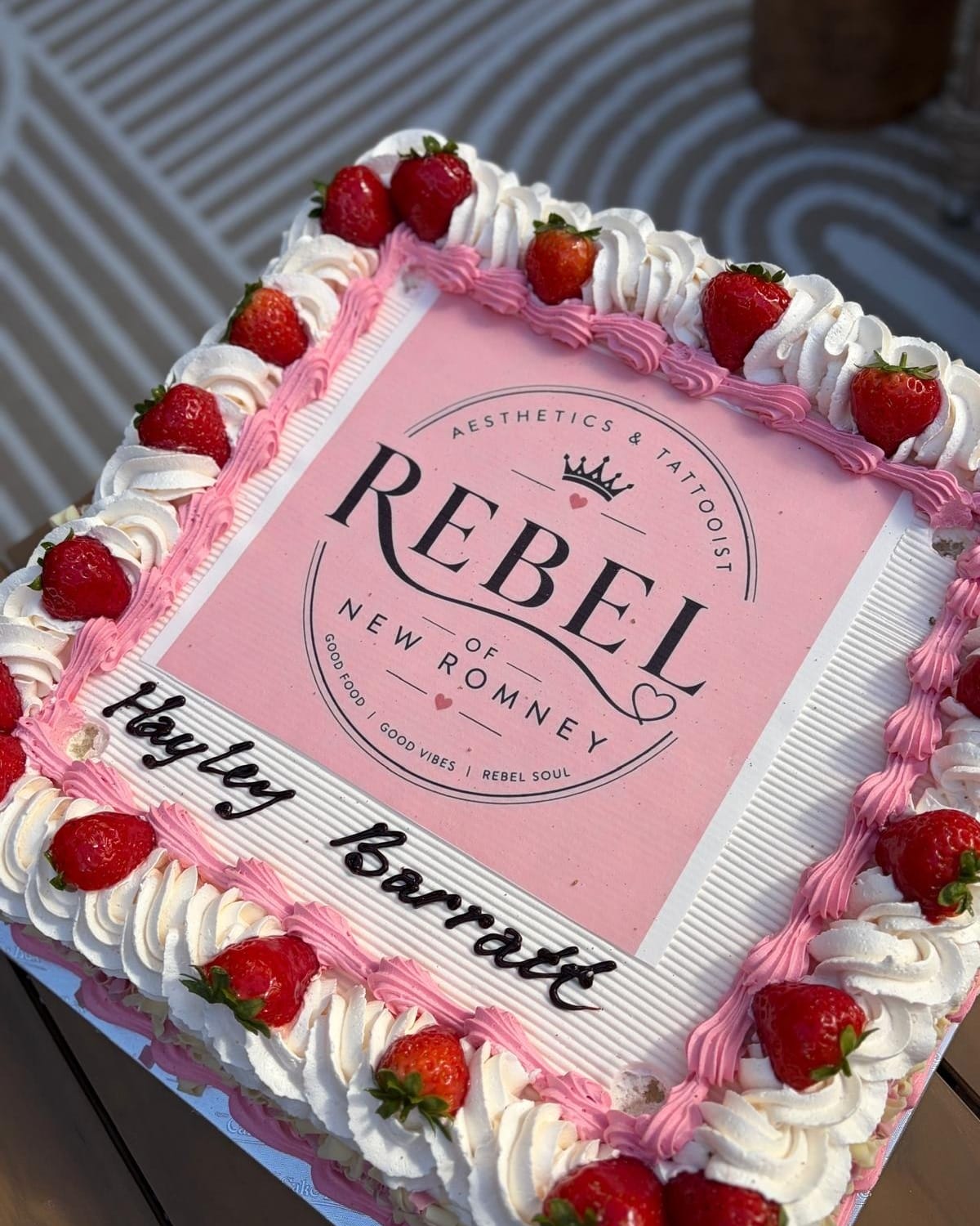 A celebration cake piped with cream and strawberries around an edible Rebel of New Romney logo