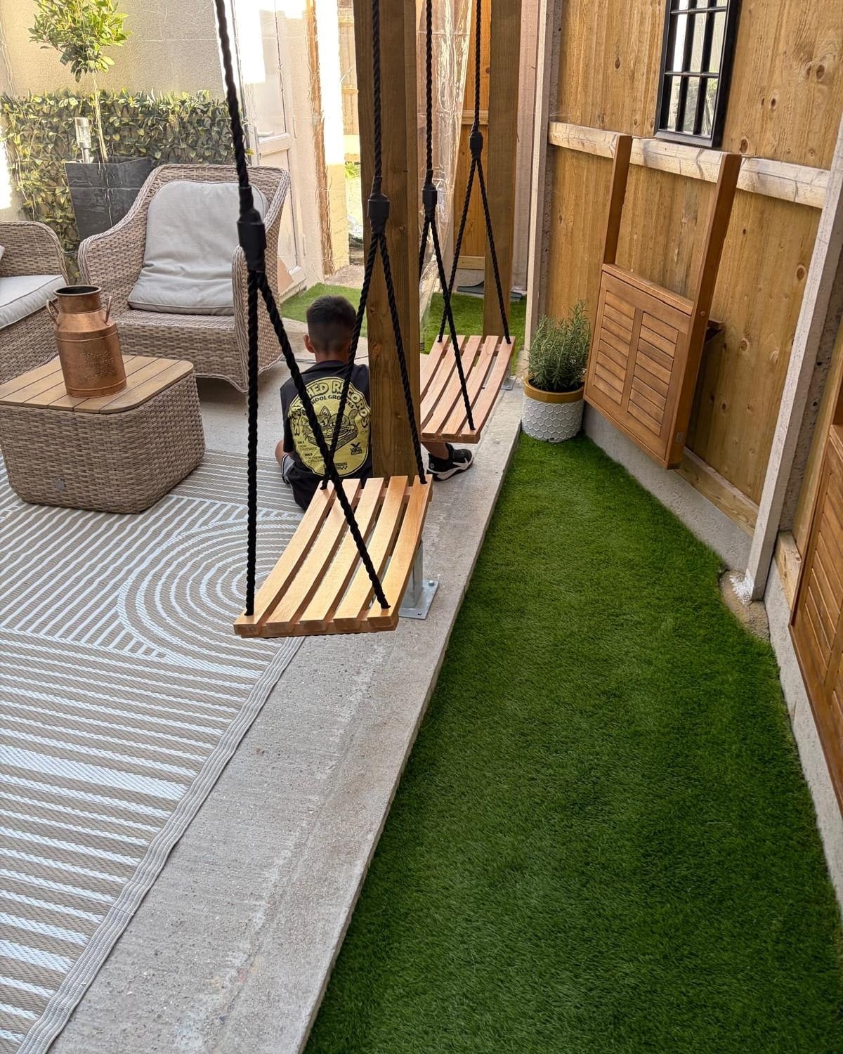Wooden swing seats hanging in the garden beside artificial grass, a rattan sofa and a copper jug on a table